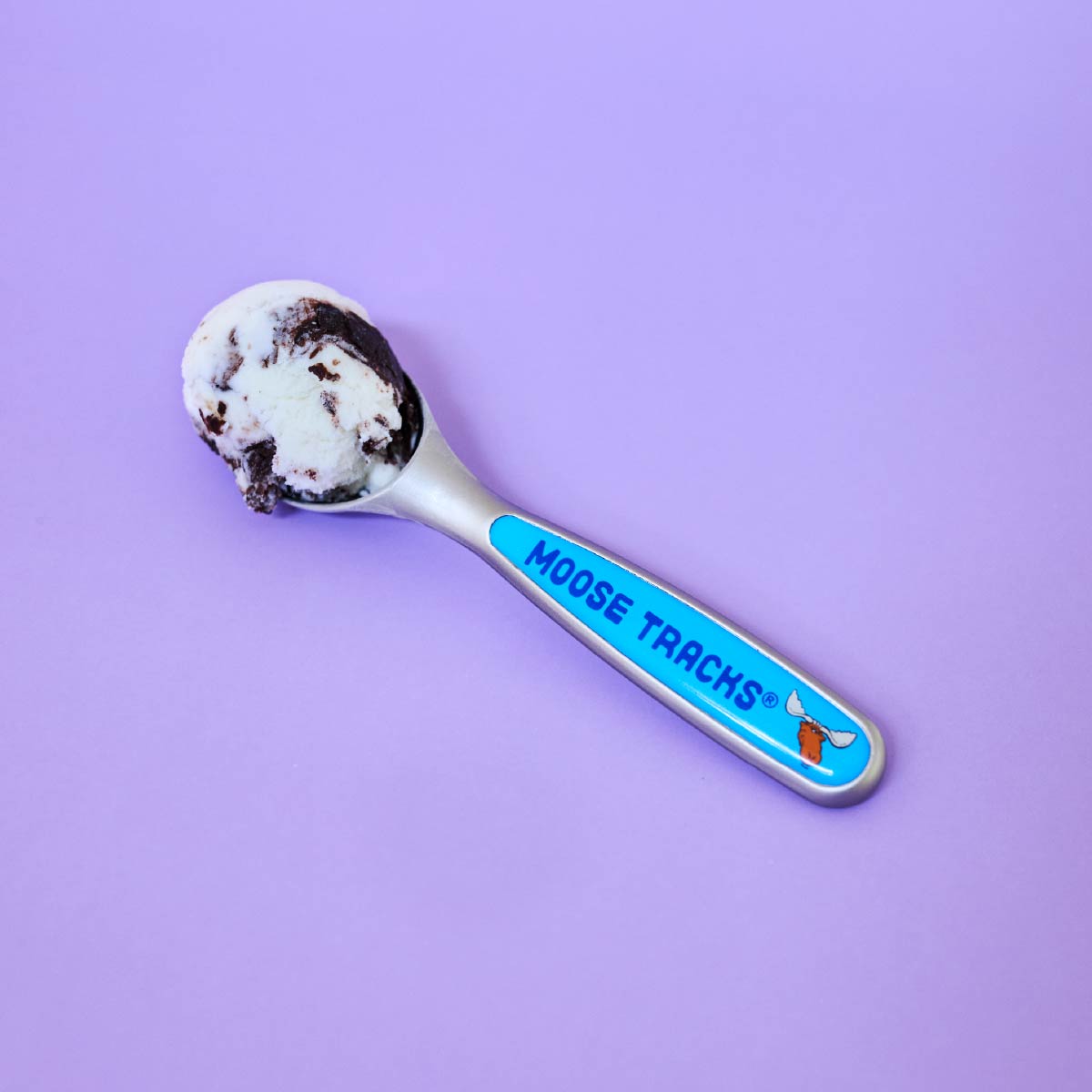 A Moose Tracks Perfectly Pint-Sized Ice Cream Scoop with a scoop of ice cream it in. The scoop has a blue design, features a moose head logo and reads, "Moose Tracks" on the handle.
