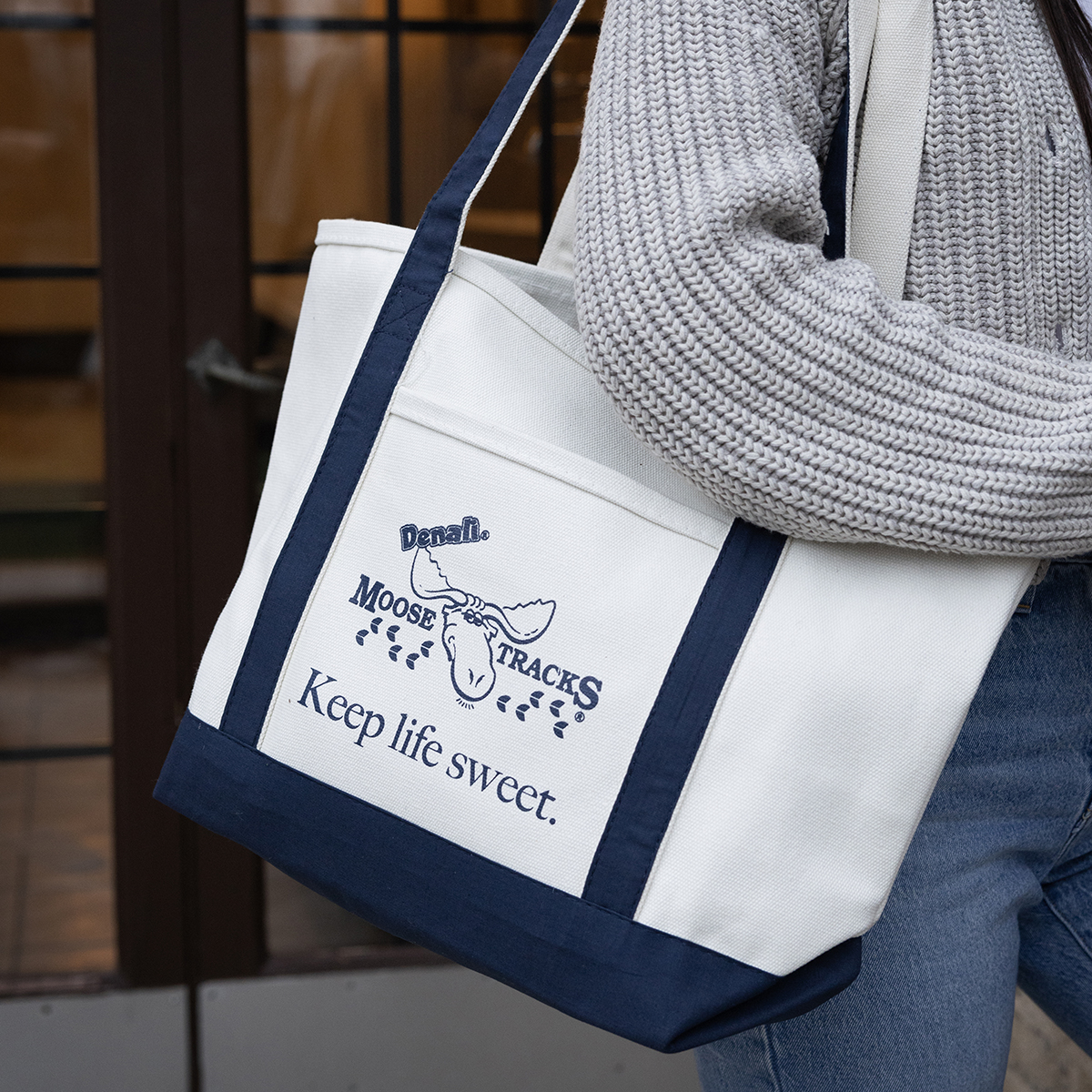 Someone carrying the Moose Tracks "Keep Life Sweet" canvas tote bag. Bag has navy detailing and a Moose Tracks logo and text reading "Keep Life Sweet" in navy-colored screen printing.