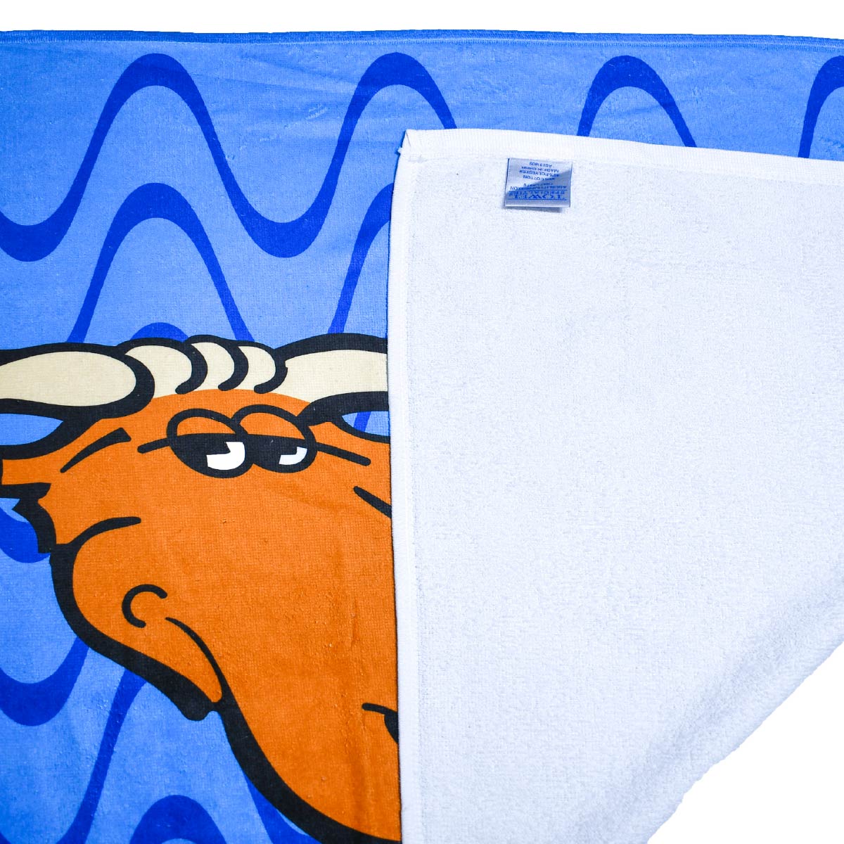 Close-up view of the Moose Tracks Beach Towel laying on a flat surface with the bottom right corner flipped over, revealing a white terry cloth underside and a sewn-in product tag. The front of the towel is bright blue with dark and light blue wavy lines in the background and features the Moose Tracks logo.