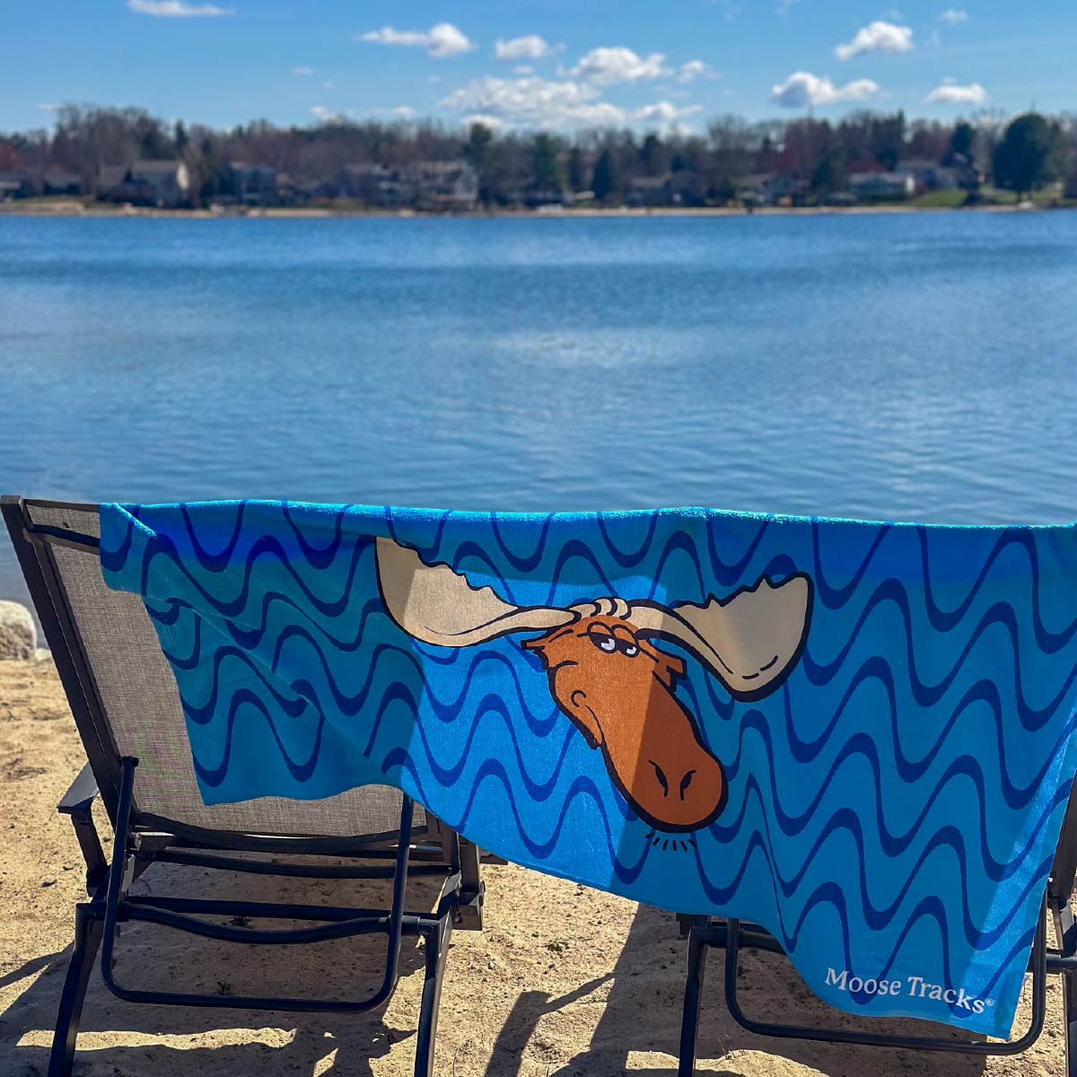 The Moose Tracks Beach Towel draped over two sun chairs with a lake in the background. The towel is bright blue with dark and light blue wavy lines in the background. The center of the towel features the Moose Tracks logo, and the bottom right corner has "Moose Tracks" printed in white.