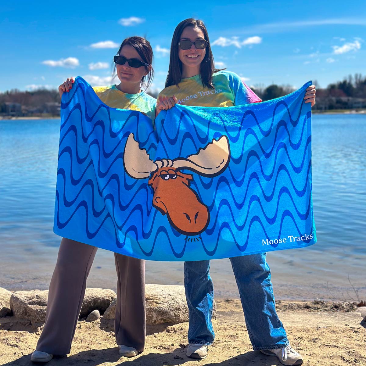 Two people holding an unfolded Moose Tracks Beach Towel with a lake in the background. The towel is bright blue with dark and light blue wavy lines in the background. The center of the towel features the Moose Tracks logo, and the bottom right corner has "Moose Tracks" printed in white.