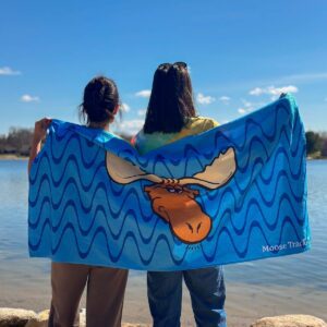 Two people with their backs turned, holding an unfolded Moose Tracks Beach Towel with a lake in the background. The towel is bright blue with dark and light blue wavy lines in the background. The center of the towel features the Moose Tracks logo, and the bottom right corner has "Moose Tracks" printed in white.