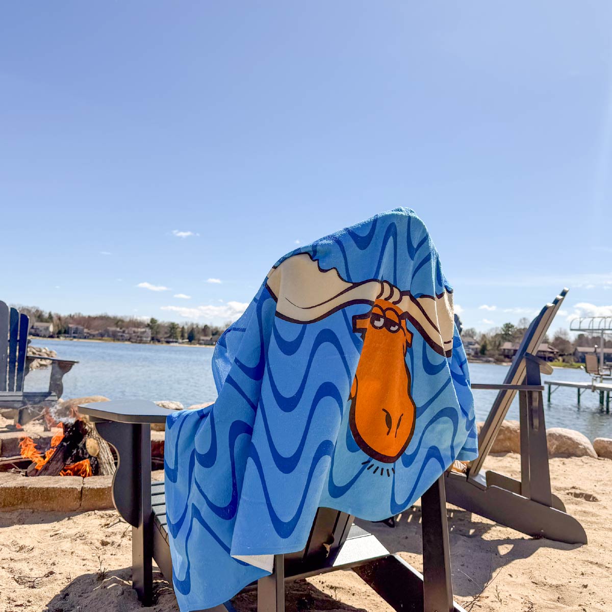 The Moose Tracks Beach Towel draped over a chair with a lake and bonfire in the background. The towel is bright blue with dark and light blue wavy lines in the background and features the Moose Tracks logo in the center of the towel.